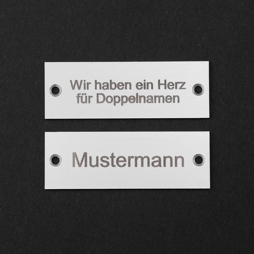 Zwei Edelstahl-Gravierschilder untereinander; auf dem unteren steht "Mustermann", auf dem oberen steht "Wir haben ein Herz für Doppelnamen"